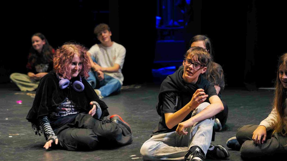 Young Company participants sit on the stage floor in Studio 2 during a rehearsal or workshop. A group of teenagers are engaged in conversation, with one person gesturing animatedly while others listen and smile.