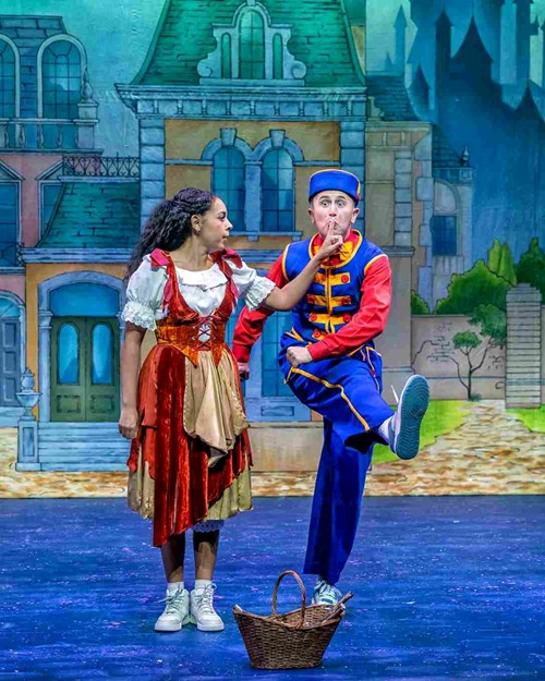 Cinderella stands next to Buttons and holds her finger to his lips. Buttons leg is raised mimicking a kick. Cinderella is wearing a brown and beige dress that looks likes rags. Buttons is wearing a blue and red bell-boy outfit.