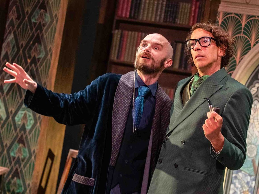 Two actors on stage in a library setting; one wearing a dark blue robe and monocle gestures dramatically while the other, in a green suit with glasses and a pipe, looks thoughtful.