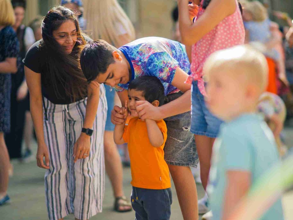 A young boy wearing an orange shirt is supported by an older child in a colourful tie-dye shirt, helping him walk. A woman with long brown hair and striped trousers bends down smiling at them. The background is filled with people enjoying an outdoor event.