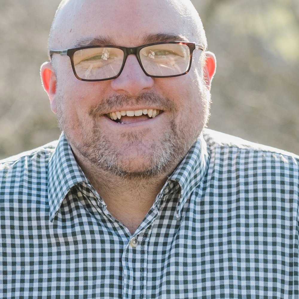 Headshot of Pete Toon. He has a shaved head, glasses, and a beard, wearing a checked shirt, smiling warmly at the camera.