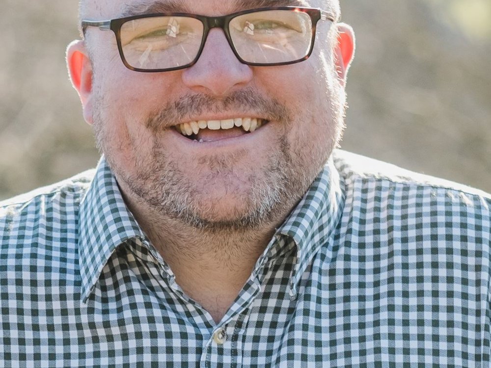 Headshot of Pete Toon. He has a shaved head, glasses, and a beard, wearing a checked shirt, smiling warmly at the camera.