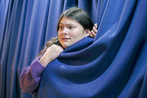 A young boy peeks nervously out from behind a thick navy curtain, gripping it tightly. He wears a purple jumper and pretends to look anxious.