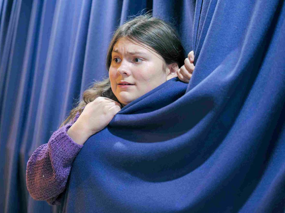 A young boy peeks nervously out from behind a thick navy curtain, gripping it tightly. He wears a purple jumper and pretends to look anxious.