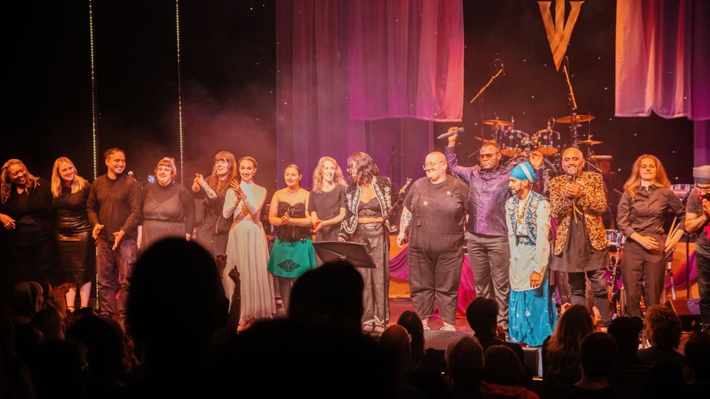 A diverse group of performers standing together on stage during a curtain call, smiling and clapping as the audience applauds. The stage is adorned with elegant drapery, lighting, and musical instruments in the background, creating a celebratory and vibrant atmosphere