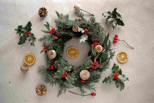 Festive Christmas wreath made of evergreen branches, holly leaves with red berries, pinecones, dried orange slices, and seed pods arranged on a neutral background, with additional holly sprigs, pinecones, and orange slices scattered around it.