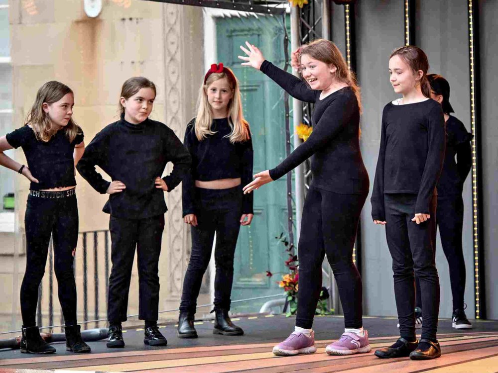 A group of six young girls perform on an outdoor stage, all dressed in black. One girl stands in the centre with arms outstretched, smiling animatedly, while the others watch her with interest. Stage lights and a decorative backdrop are visible.