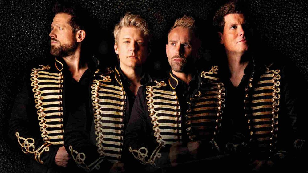 Four men wearing black jackets with ornate gold detailing stand against a dark background, looking in different directions with confident, dramatic expressions.