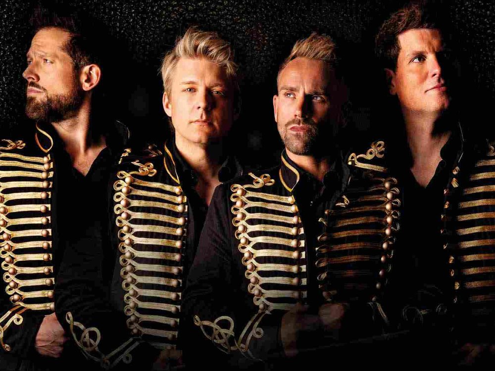 Four men wearing black jackets with ornate gold detailing stand against a dark background, looking in different directions with confident, dramatic expressions.