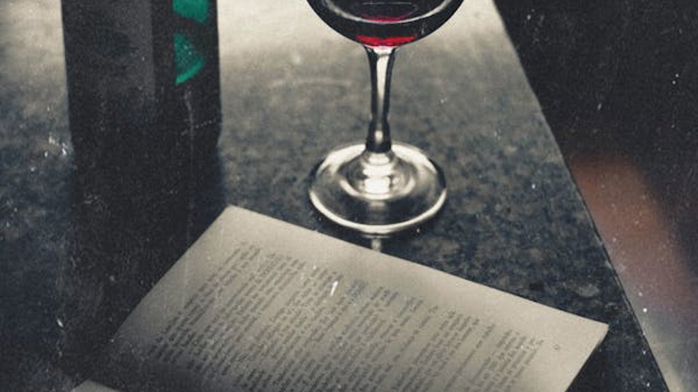 The image shows a vintage scene with a partially opened book on a dark surface, its pages reflecting the light. Beside the book is a wine glass filled with dark red wine, with a green wine bottle placed behind it. The atmosphere has a moody, nostalgic feel, with soft lighting accentuating the textures of the objects.