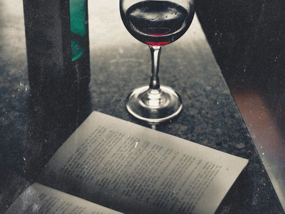 The image shows a vintage scene with a partially opened book on a dark surface, its pages reflecting the light. Beside the book is a wine glass filled with dark red wine, with a green wine bottle placed behind it. The atmosphere has a moody, nostalgic feel, with soft lighting accentuating the textures of the objects.