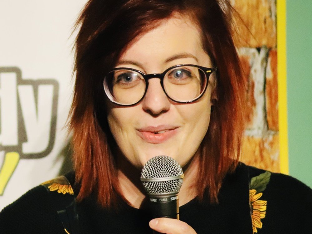 A woman with short red hair and round glasses speaks into a microphone onstage, smiling slightly. She wears a black top with sunflower-patterned straps, and a comedy club backdrop is visible behind her