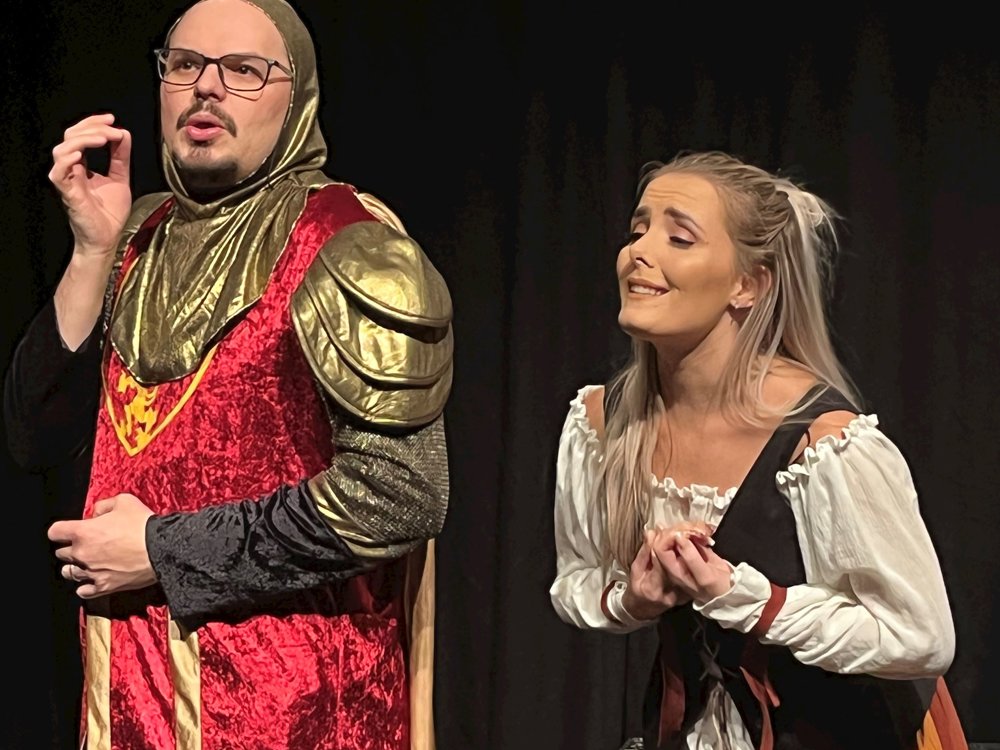 A performer in a red and gold costume gestures theatrically while another actor watches, smiling in a humorous stage moment.