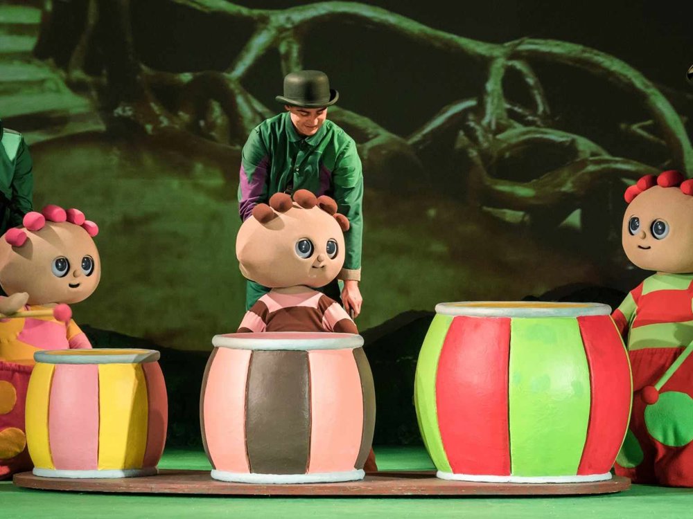 The Tombliboos play large, brightly coloured drums on stage, with performers in green costumes assisting them in a woodland scene.