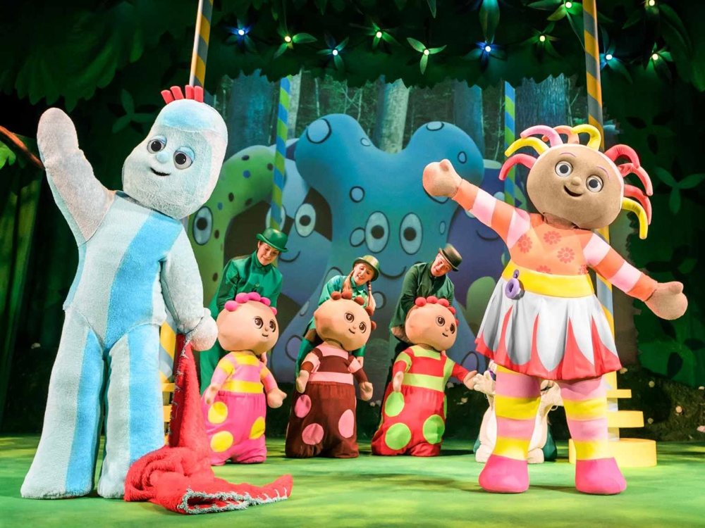 Igglepiggle and Upsy Daisy wave to the audience alongside the Tombliboos, with performers dancing behind them in a vibrant forest set.