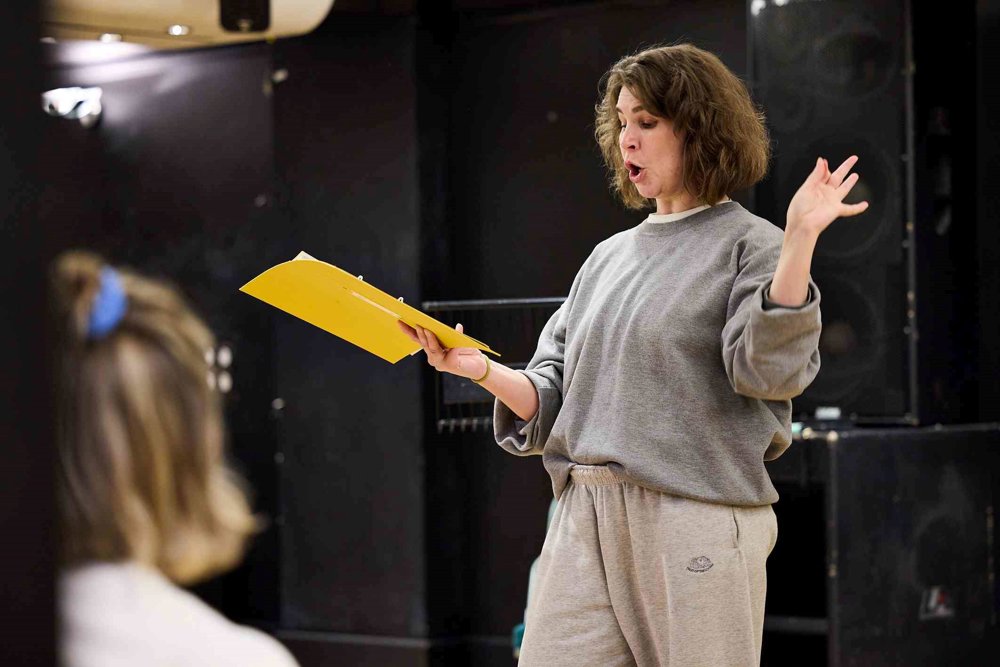 An actor in a grey sweatshirt and matching trousers performs passionately during a rehearsal, holding a yellow folder in one hand and gesturing expressively with the other.