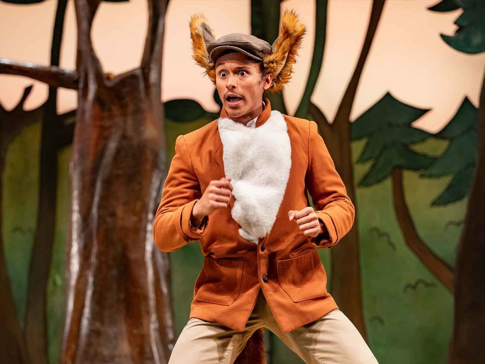 An actor dressed as a fox stands alert on a stage set resembling a forest, with wide eyes and raised hands as if startled or cautious.