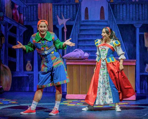 Two performers are on stage in a vibrant and colourful setting. The character on the left wears a whimsical, oversized outfit featuring a green shirt, striped overalls, a bright orange backwards cap, and red sneakers, with an expressive, comedic pose and exaggerated facial expression. The character on the right is dressed as a princess, wearing a bright costume with a red and blue bodice, puffy white sleeves, and a skirt decorated with floral patterns, paired with white sneakers.