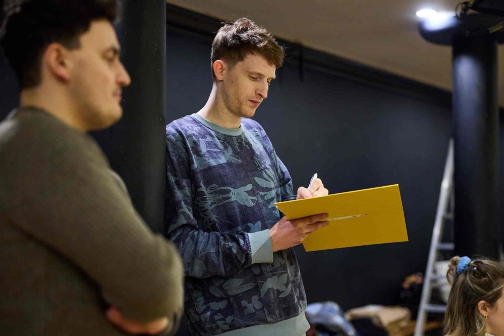 A man in a patterned sweatshirt leans against a black pillar, focused on writing in a yellow folder. Another person is blurred in the foreground, and rehearsal props are visible in the background.