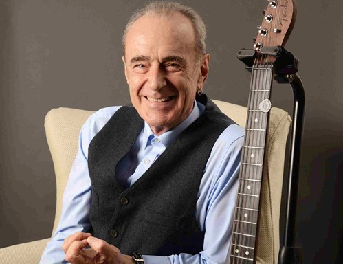 Francis Rossi sits in a chair next to a guitar on a stand and smiles at the camera. 