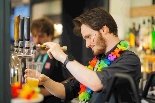 A Lawrence Batley Theatre team member pours a drink at the bar, wearing a colourful flower garland and face paint, with a focused expression. The bar is lively and well-stocked, with another staff member visible in the background.