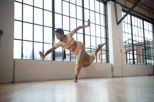 A South Asian classical dancer balances gracefully on one foot in a contemporary studio space with tall industrial windows. She wears a gold traditional outfit and performs a dynamic movement with arms extended and one leg lifted behind her.