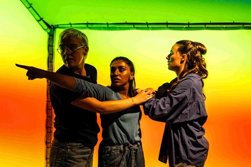 Against a background of green, yellow and orange colours stand three young people. The middle person is pointing off into the distance with the other two people trying to hold them back.  