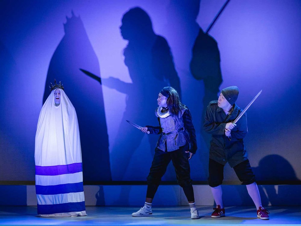 An image of a blue-lit stage, two actors holding knives confront a ghostly crowned figure in white robes, whose enlarged shadow looms behind them.