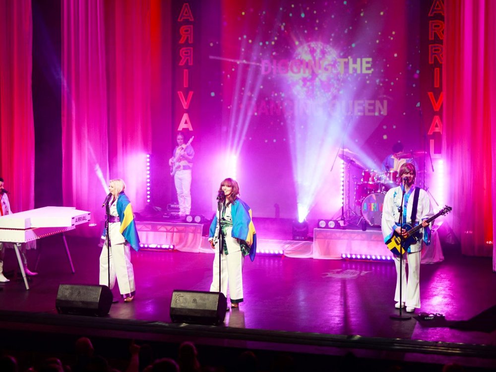 The full band performs on stage in ABBA-inspired costumes beneath vibrant pink lighting, with a disco-themed backdrop and the words "Dancing Queen" displayed on the screen.
