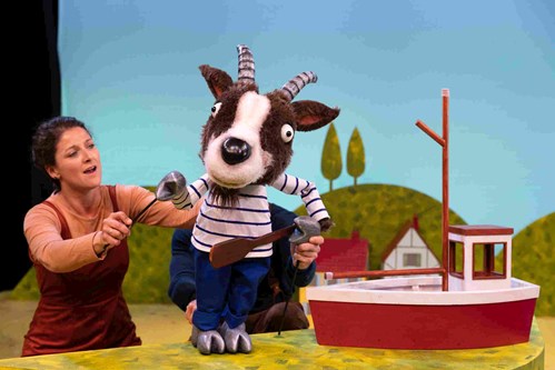 A goat puppet dressed in blue trousers and a white and blue striped t-shirt holds an oar as he stands next to a boat. 