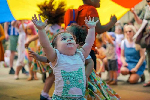A small child with a cat design on her white shirt reaches up excitedly towards the sky under a large, multicoloured parachute. Other children and adults are engaged in the festivities in the background.