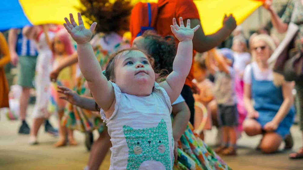 A small child with a cat design on her white shirt reaches up excitedly towards the sky under a large, multicoloured parachute. Other children and adults are engaged in the festivities in the background.