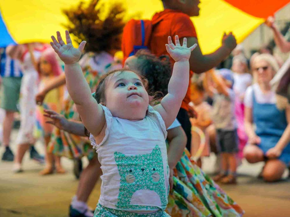 A small child with a cat design on her white shirt reaches up excitedly towards the sky under a large, multicoloured parachute. Other children and adults are engaged in the festivities in the background.