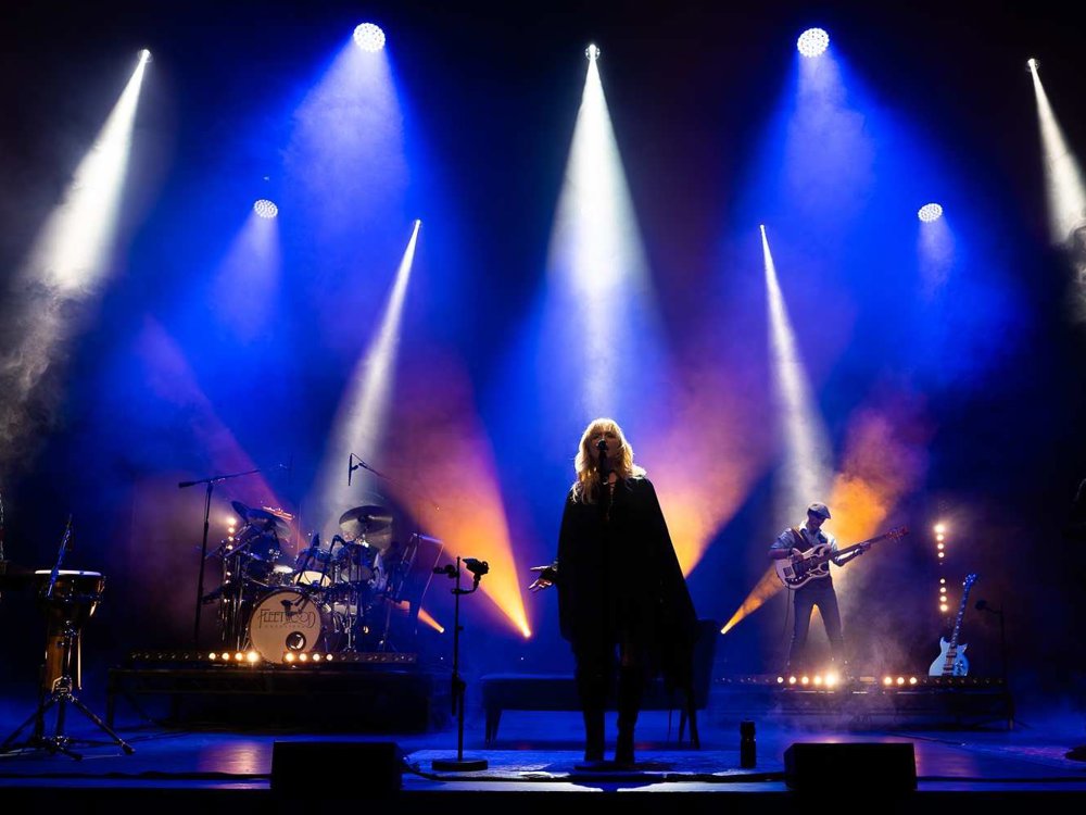 A full band performs on stage under dramatic white, blue, and orange spotlights, with musicians spaced across the stage playing keyboard, drums, guitar, and bass as smoke rises around them.