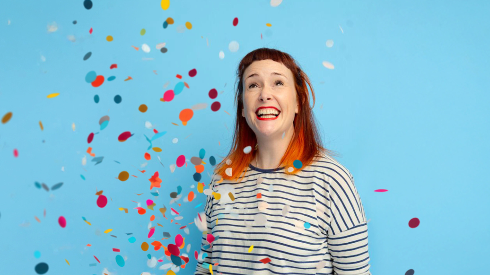 Smiling red-haired woman in a striped top looks upward as colourful confetti falls against a bright blue background.