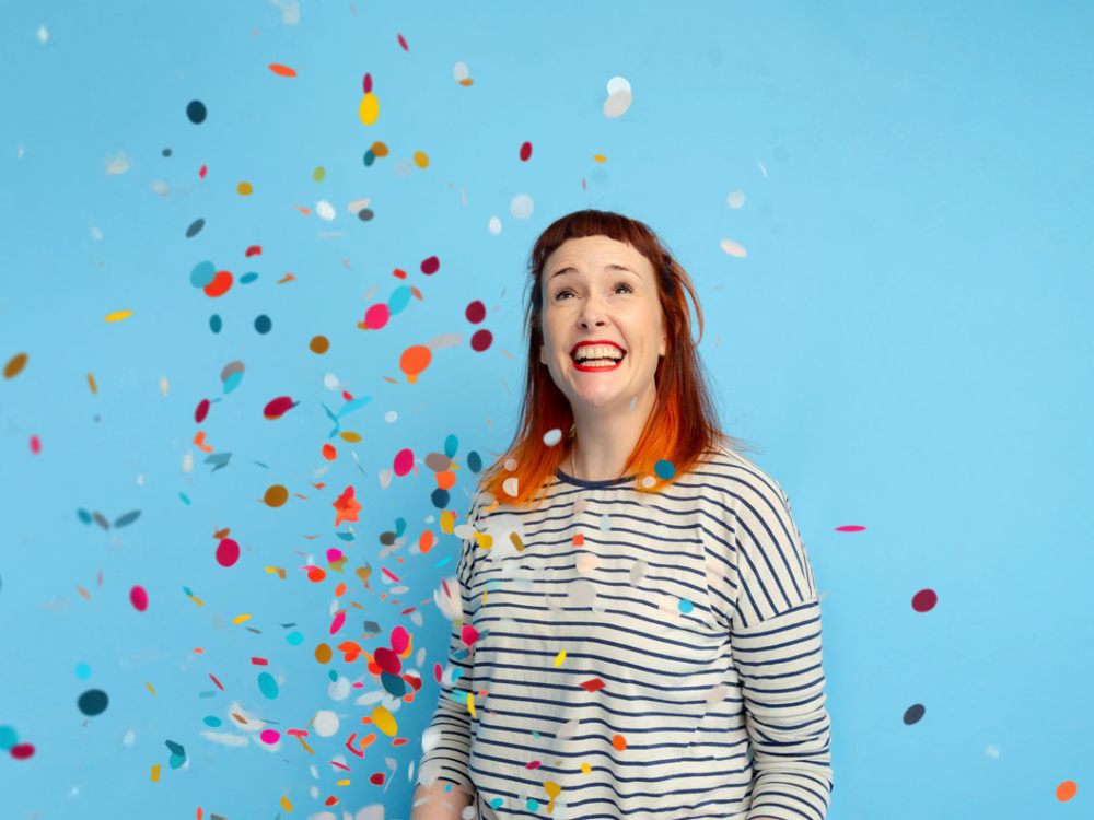 Smiling red-haired woman in a striped top looks upward as colourful confetti falls against a bright blue background.