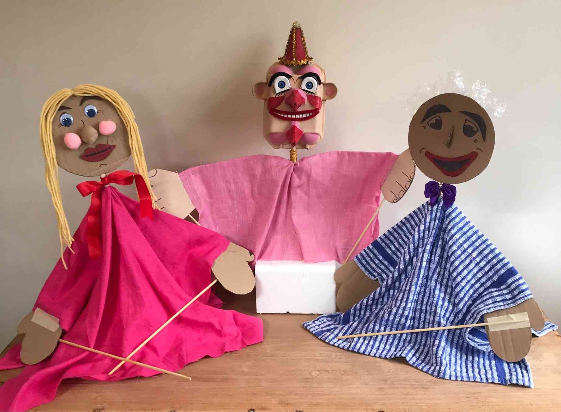 Three handmade puppets displayed on a wooden table against a plain background. Two larger puppets wear colourful fabric costumes — one in bright pink with blonde yarn hair, the other in blue striped fabric with white hair — while a smaller clown-like puppet in pink stands between them. The puppets are made from cardboard, fabric and craft materials, with wooden sticks attached for movement.