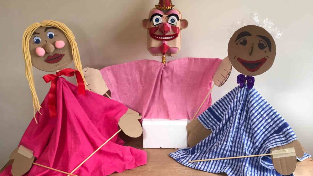 Three handmade puppets displayed on a wooden table against a plain background. Two larger puppets wear colourful fabric costumes — one in bright pink with blonde yarn hair, the other in blue striped fabric with white hair — while a smaller clown-like puppet in pink stands between them. The puppets are made from cardboard, fabric and craft materials, with wooden sticks attached for movement.