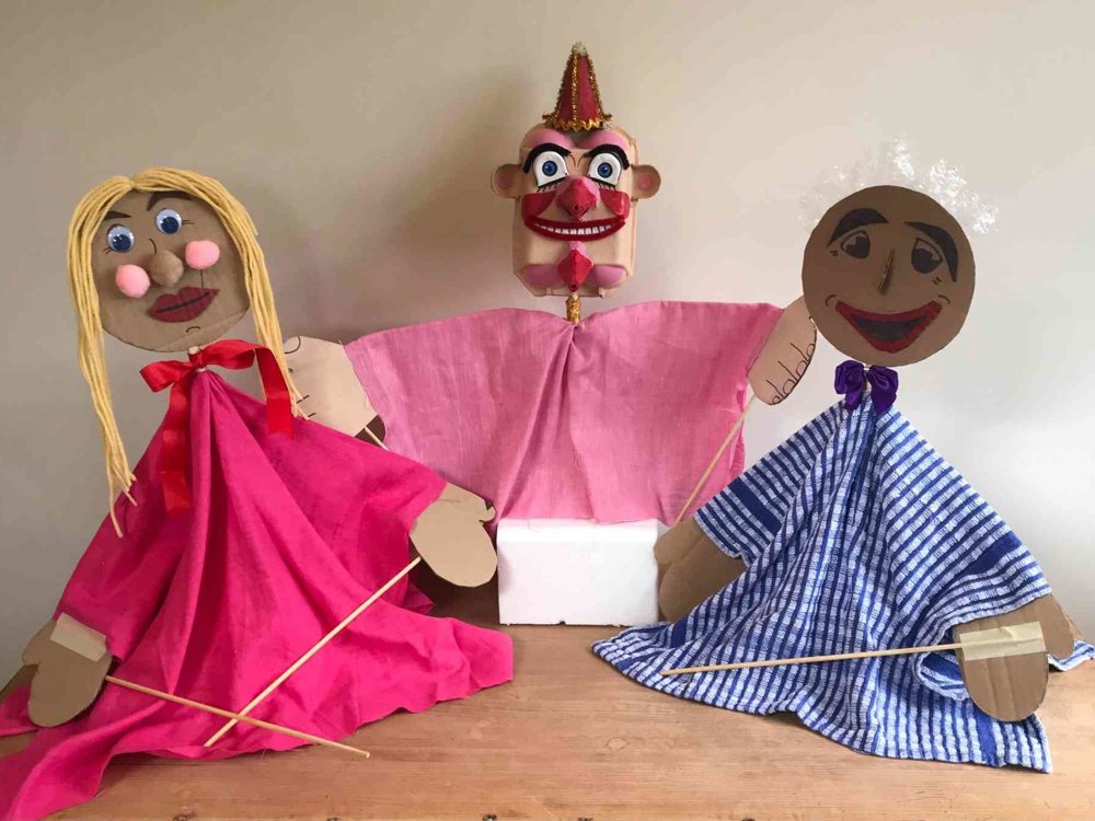 Three handmade puppets displayed on a wooden table against a plain background. Two larger puppets wear colourful fabric costumes — one in bright pink with blonde yarn hair, the other in blue striped fabric with white hair — while a smaller clown-like puppet in pink stands between them. The puppets are made from cardboard, fabric and craft materials, with wooden sticks attached for movement.
