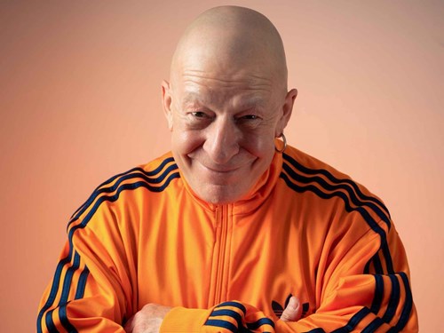 Comedian Jeff Innocent is grinning, he is wearing a bright organge tracksuit jacket. 