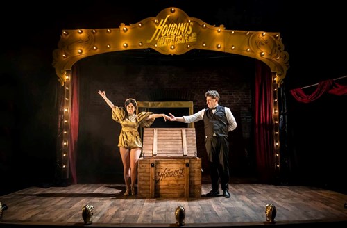 There is a large, wooden box in the centre of the stage. Bess and Houdini stand on either side of the box, their arms are outstretched and they hold hands. 