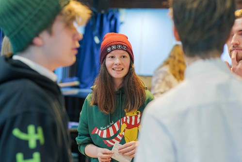 A young person wearing a red beanie and a green Winnie the Pooh hoodie stands smiling among a small group, engaged in conversation.