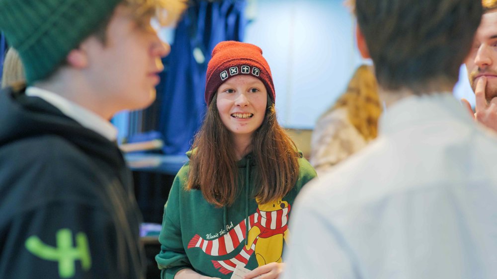 A young person wearing a red beanie and a green Winnie the Pooh hoodie stands smiling among a small group, engaged in conversation. 