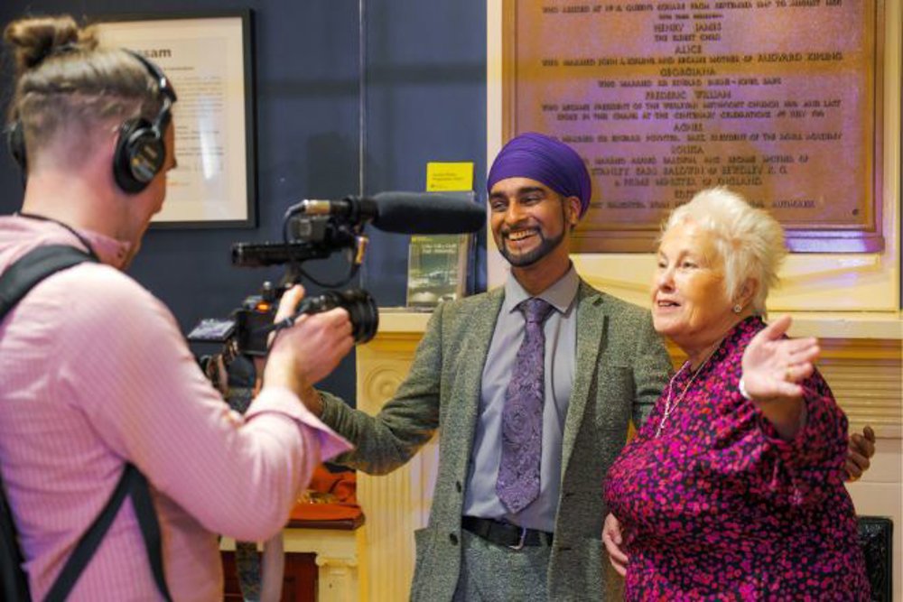 A man wearing a purple turban and a grey suit smiles warmly while standing next to an older woman in a pink and black patterned dress. They are being filmed by a videographer in a pink shirt holding a camera and microphone, set against an indoor backdrop with framed documents and a wooden plaque.