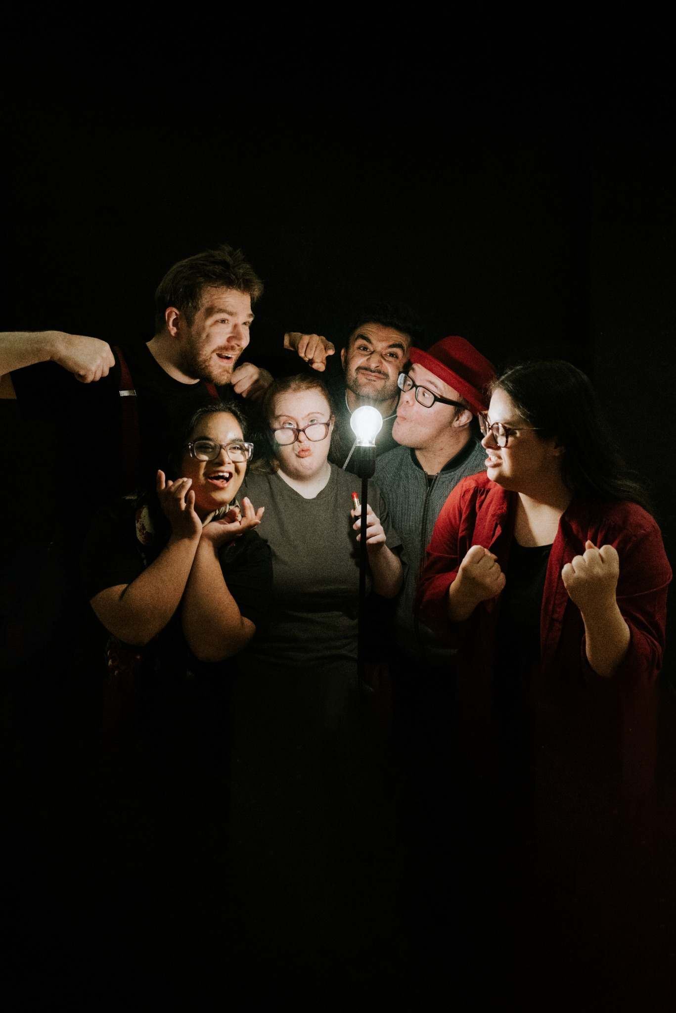 Six people stand closely together against a dark background, playfully posing around a glowing light bulb on a stand. They pull exaggerated expressions and gestures, creating a lively, humorous group portrait with theatrical energy.