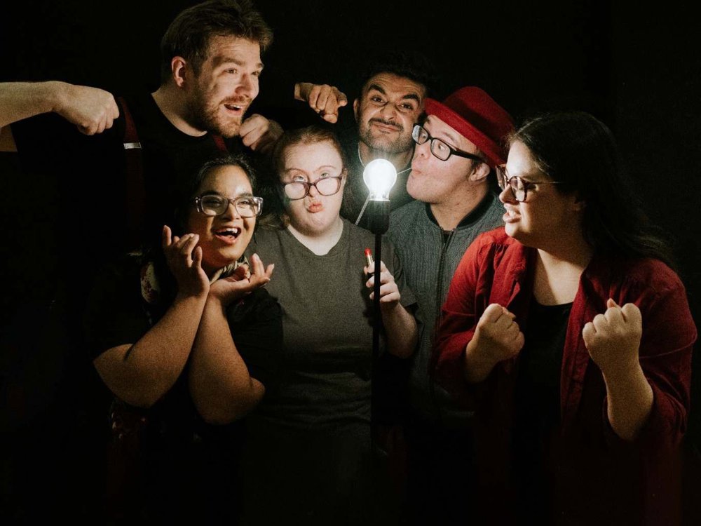 Six people stand closely together against a dark background, playfully posing around a glowing light bulb on a stand. They pull exaggerated expressions and gestures, creating a lively, humorous group portrait with theatrical energy.