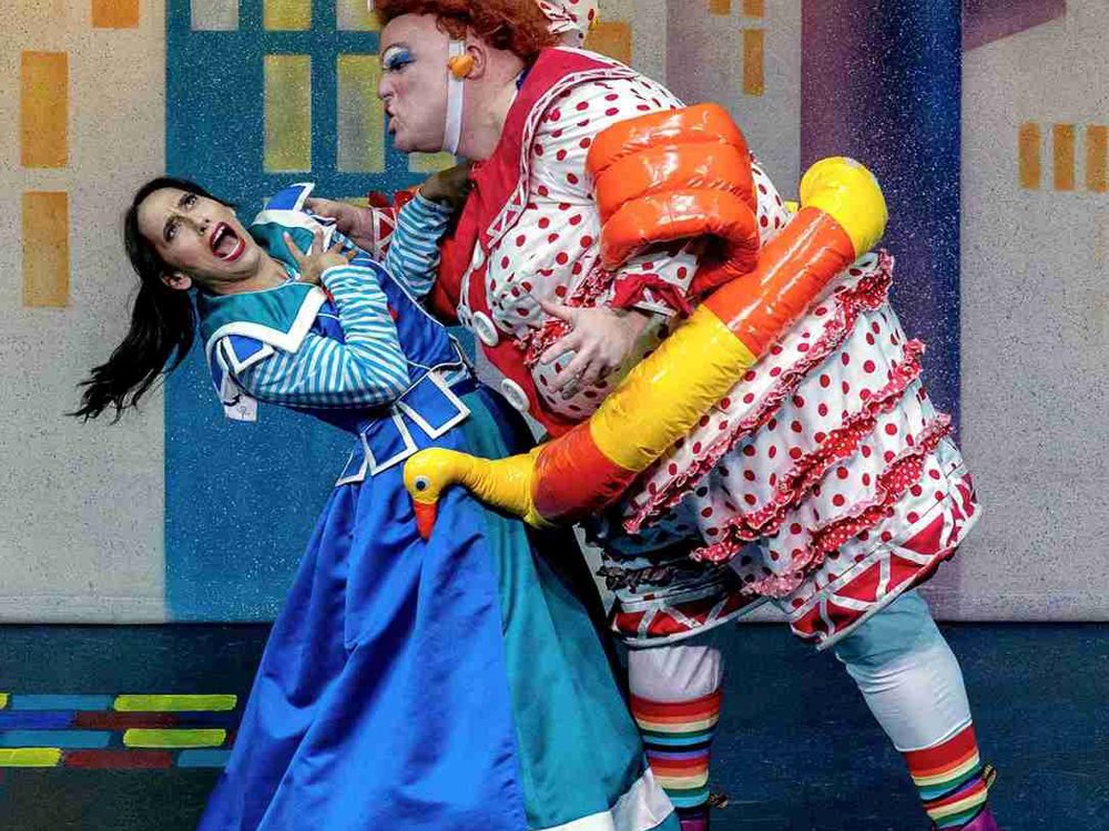 A vibrant and comedic stage scene featuring two characters in exaggerated costumes. On the left, a woman in a bright blue dress with white trim and a sailor-style collar expresses mock fear, her mouth open in a dramatic scream. On the right, a larger-than-life character in an elaborate polka-dot and ruffled outfit leans toward her with a playful, exaggerated scowl. 