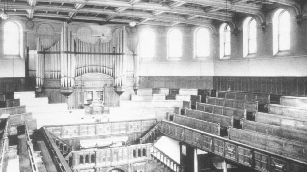A black-and-white photograph of the theatre when it was a mission. At the rear there is a large organ and there are pews/benches around the four corners of the building.