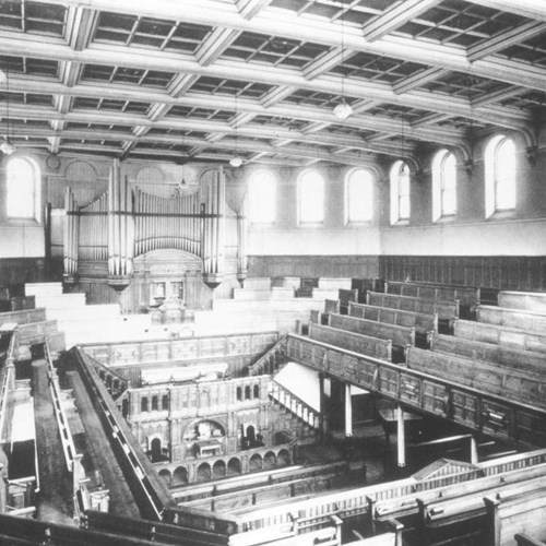 A black-and-white photograph of the theatre when it was a mission. At the rear there is a large organ and there are pews/benches around the four corners of the building.