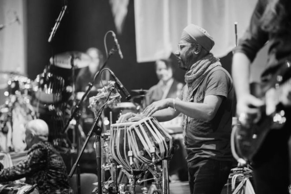 A black-and-white image of a musician playing the tabla on a stage during a live performance. The musician, wearing a turban and scarf, is surrounded by other performers and instruments, creating a dynamic and engaging scene with a focus on traditional music